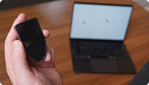 Hand holding a black smartphone-like device in front of an open laptop on a wooden table.